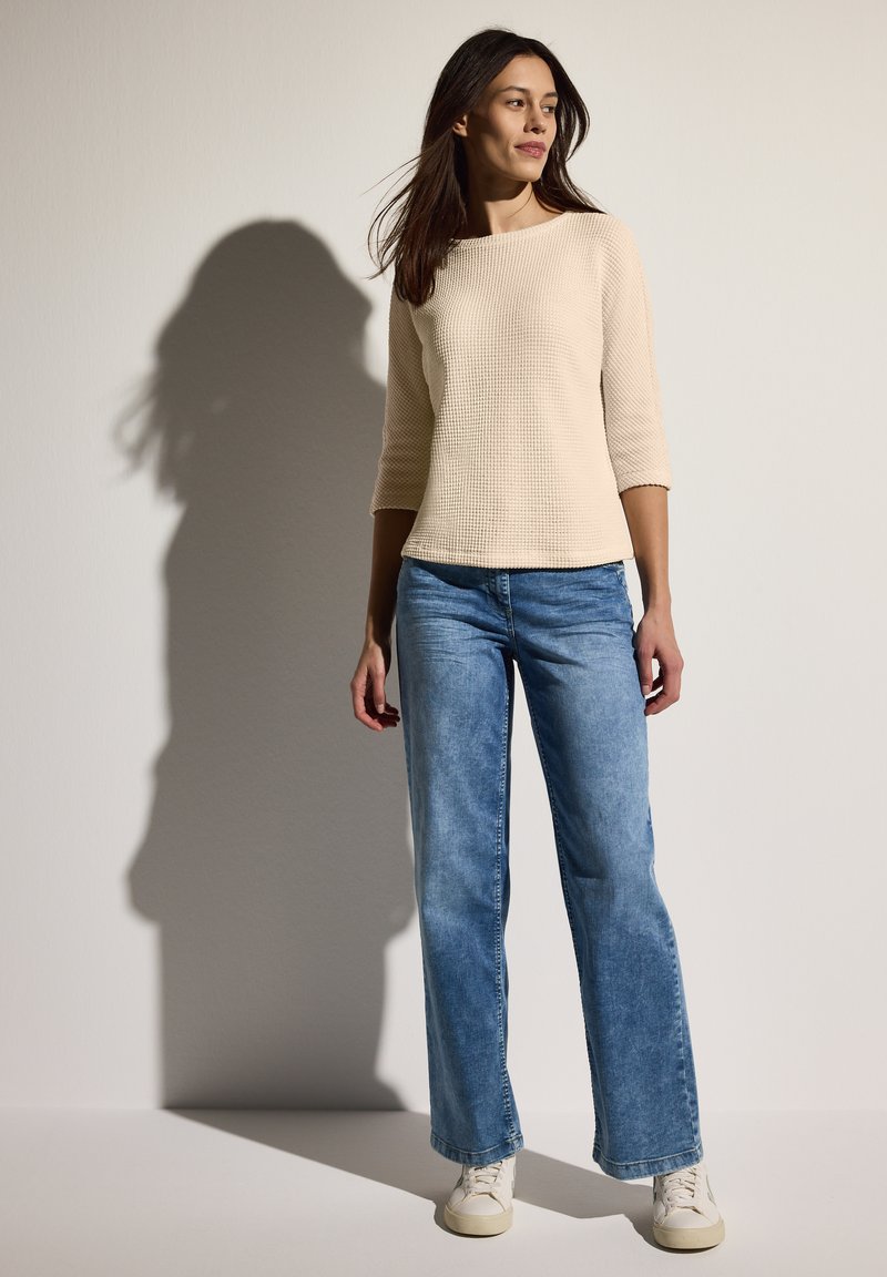 Beige strukturierter Langarm-Top, kombiniert mit hellblauen weit geschnittenen Jeans und weißen Sneakers. Einfaches Design, keine sichtbaren Muster.