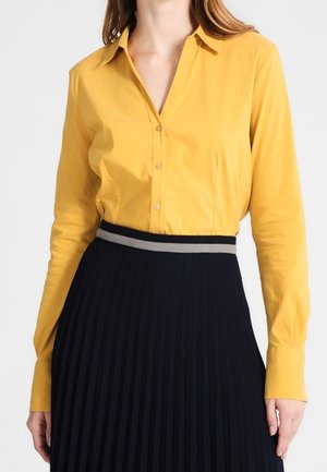 Vrouw draagt een mosterdgele blouse met knopen, ingestopt in een zwarte geplooide rok met hoge taille en een grijze tailleband.