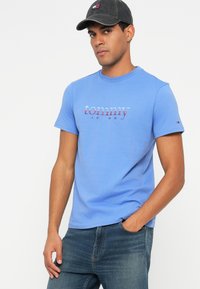 Modra bombažna majica s "tommy jeans" logotipom v rdeči in temno modri barvi, v kombinaciji s temnimi kavbojkami in temnim klobukom z logotipom.