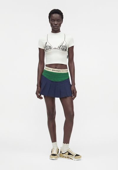 Γυναίκα που φοράει λευκό cropped t-shirt με γραφικό σουτιέν, πράσινη και ναυτικό πλισέ φούστα, λευκές κάλτσες και μπεζ και μαύρα αθλητικά παπούτσια, όρθια.