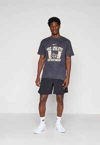 Must Nike spordisärk valge trükitud logoga, koos mustade lühikestega. Modell kannab valgeid tenniseid ja pahkluu sokke.