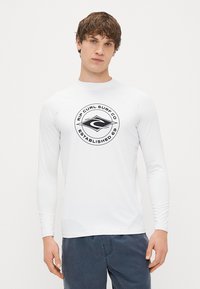 Λευκό πουκάμισο σερφ με μακριά μανίκια, με μαύρο κυκλικό λογότυπο που περιλαμβάνει την ένδειξη "Rip Curl Surf Co" και "Established 69" τυπωμένα στο μπροστινό μέρος.