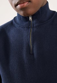 Pull à fermeture éclair bleu marine avec col montant et texture en maille fine. Dispose d'une fermeture éclair argentée et d'accents côtelés au col et à l'ourlet.