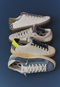 Quatre baskets décontractées en blanc, beige et bleu avec des lacets, des logos de marque et des accents colorés au talon disposées sur un fond bleu.