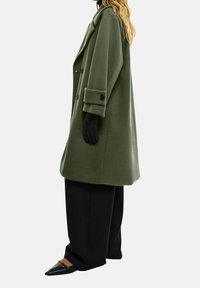 Grøn uldblandingsfrakke med en afslappet pasform, dobbeltbrystet design, stor krave og sorte knapdetaljer, parret med løse sorte bukser og sorte sko.