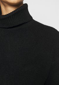 Gros plan sur une personne portant un pull en tissu côtelé noir avec un col roulé sur un fond uni.