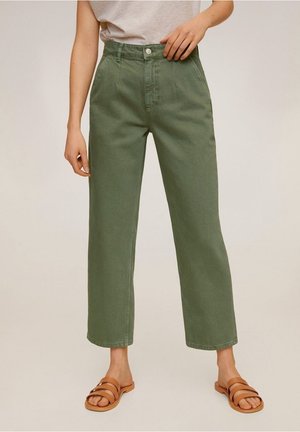 Persoon draagt olijfgroene, rechte broek met hoge taille, beige top en bruine sandalen met dubbele band en platte zool, staat met één hand bij de taille.