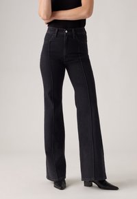 Mujer que lleva vaqueros de campana negros de cintura alta con detalle de costura frontal, combinados con botas de tacón negro de punta afilada y una blusa sin mangas negra.