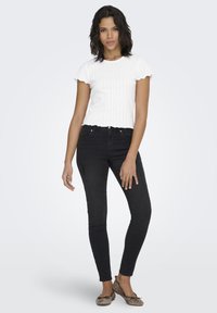 Wit geribd kortermig shirt met geschulpte randen, gecombineerd met zwarte skinny jeans en lichtbruine ballerina's. Model staat met één hand op de heup.