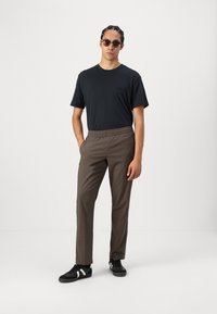 Svart t-skjorte, brune bukser med elastisk midje, svarte joggesko med hvite detaljer. Modell som har på seg solbriller, står mot en ensfarget bakgrunn.