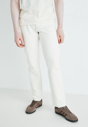 Jeans bianchi in cotone con una vestibilità slim, abbinati a sneaker in suede marrone con lacci scuri e strisce a tre tonalità. Sfondo bianco.