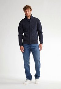 Pulover tricotat bleumarin cu fermoar, guler și manșete cu dungi, asortat cu blugi bleu deschis și adidași albi. Design simplu și texturat.