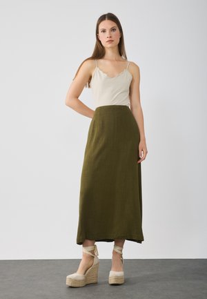 Camisole beige clair avec des bords festonnés, associée à une longue jupe verte olive, complétée par des espadrilles de couleur claire.