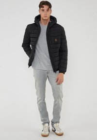 Giacca a vento nera con cappuccio, texture trapuntata, chiusura con zip e tasca sul petto. Indossata sopra una maglietta grigia e jeans chiari con sneakers.