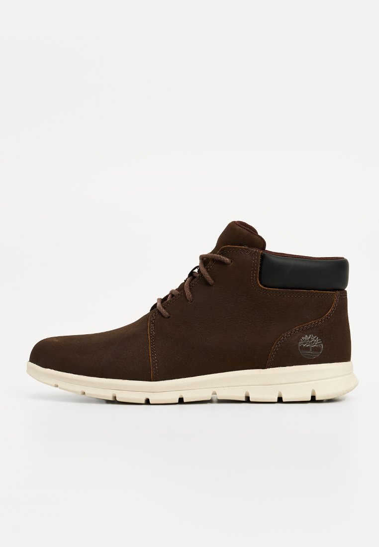 Botas de cuero marrón hasta el tobillo con una suela de goma blanca, detalles de costura y un acento negro en el cuello. Presenta un logotipo en el lateral.