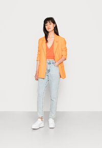 Blazer laranja sobre uma blusa vermelha, combinado com jeans de cintura alta de tom azul-claro e ténis brancos. O conjunto apresenta elementos de design minimalistas.
