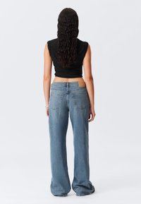Woman with long dark wavy hair wearing a sleeveless black top and loose blue jeans, standing facing away against a plain background.