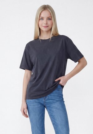 Junge Frau mit langen blonden Haaren, die ein lockeres dunkelgraues T-Shirt und blaue Jeans trägt, steht vor einem schlichten hellen Hintergrund.
