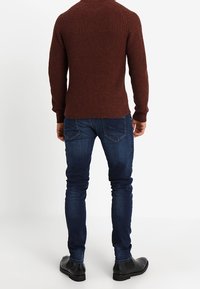 Brun, ribbad stickad tröja med långa ärmar, kombinerad med mörkblå jeans och svarta ankelskor. Sedd bakifrån.