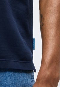 Κοντινό πλάνο ενός ατόμου που φοράει μπλε πουκάμισο και μπλε τζιν, με μια μικρή μπλε υφασμάτινη ετικέτα στη ραφή της πλευράς του πουκαμίσου.