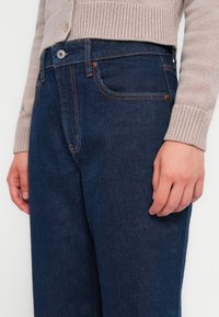 Jeans de mezclilla azul oscuro con cintura alta, corte casual y costuras naranjas contrastantes. Cuenta con bolsillos laterales y cierre de botón metálico.