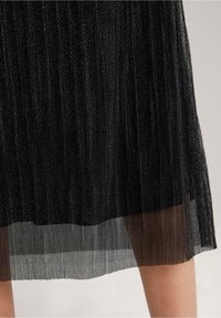 Černá plisovaná sukně s lesklou texturou. Má vrstvený design s průhlednou látkou na spodním okraji, který vytváří lehký, plynulý vzhled.