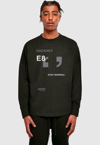 Must musta särk, millel on pikad varrukad, valmistatud puuvillast, esiküljel on graafiline tekst "HACKNEY E8" ja "STAY SURREAL". Omab mugavat lõiget ja ümmargust kaelust.