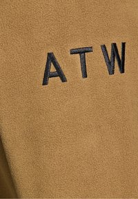 Tan suede fabric with a soft texture. Features black embroidered letters "ATW" prominently displayed. Close-up view shows fine detail.