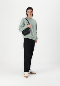 Tricou sudat de culoare verde deschis, pantaloni negri și adidași bej cu dungi negre. Modelul poartă o geantă mică, neagră, purtată pe umăr.