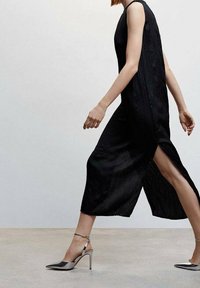 Vestido negro sin mangas con acabado texturizado y aberturas laterales. Combinado con zapatos de tacón alto de patente plateados con correas en los tobillos.