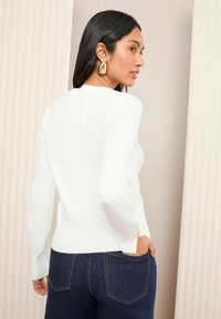 Weißer Langarmpullover mit geripptem Saum und rundem Halsausschnitt, kombiniert mit dunkelblauen Jeans. Hat eine figurbetonende Silhouette und eine glatte Textur.