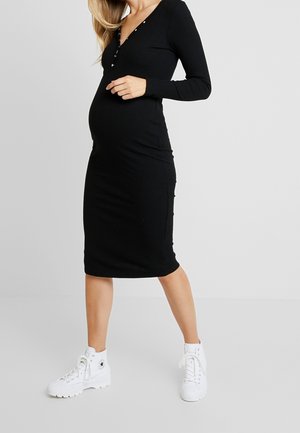 Gravid kvinde iført en tætsiddende sort langærmet kjole og hvide høje sneakers står foran en ensfarvet grå baggrund.