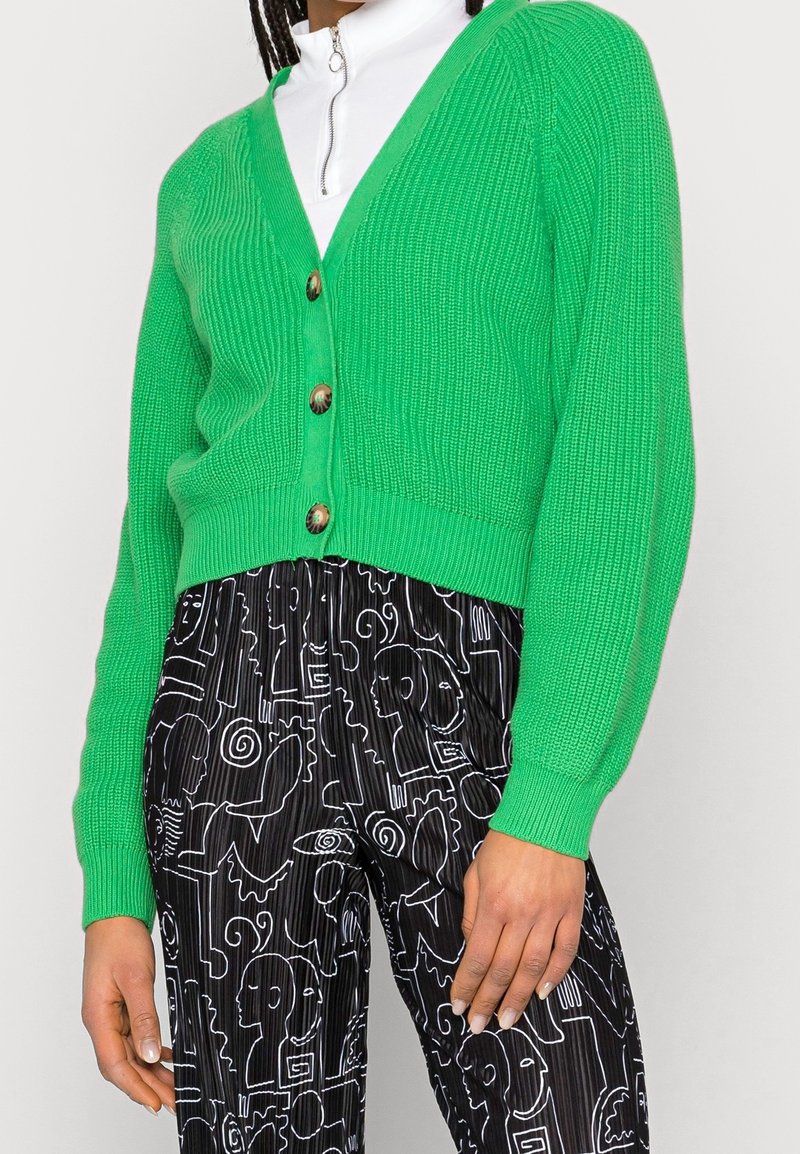 Grön ribbad knappad cardigan över en vit zip-up-tröja, kombinerad med svarta byxor med vita abstrakta linjemönster.