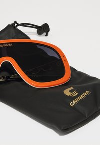 Occhiali da sole arancioni con una grande montatura e lenti scure, appoggiati su un astuccio in pelle nera con il logo dorato Carrera.