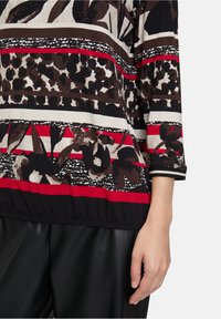 Top a fantasia mista di nero, marrone e crema con strisce rosse, disegni floreali e orlo arricciato. Maniche lunghe con polsini a contrasto.