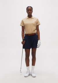 Γυναίκα που φοράει μπεζ πόλο και σκούρο μπλε σορτς κρατάει μπαστούνι του γκολφ στο δεξί χέρι και φοράει λευκό γάντι, κάλτσες και παπούτσια μπροστά σε απλό φόντο.