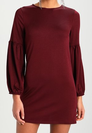 Femme portant une robe bordeaux à manches longues bouffantes froncées et col rond, debout devant un fond uni.