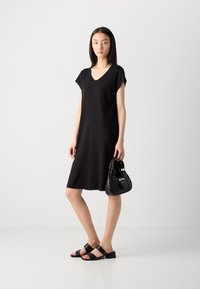 Black, knee-length dress with a V-neck and short sleeves, paired with a small black handbag and black cross-strap sandals. Smooth texture.