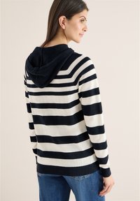 Femme de profil portant un sweat à capuche à rayures noires et blanches et un jean bleu sur un fond clair uni.