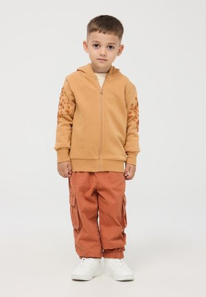 Felpa con cappuccio e zip di colore marrone chiaro, con dettagli testurizzati sulle maniche, abbinata a pantaloni cargo arancioni dal taglio comodo e scarpe da ginnastica bianche.