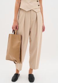 Pantaloni beige su misura con pieghe frontali, abbinati a una borsa a tracolla color cammello. Muli neri dal design semplice completano l'outfit.