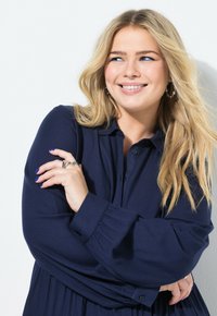 Marineblauwe blouse met een knoopsluiting aan de voorkant, zachte stof en gerimpelde mouwen. Lange, vloeiende snit met een gladde textuur en subtiele glans.