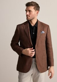 Brauner Leinenblazer mit Reverskragen, zwei Fronttaschen und einem hellblauen Taschentuch, kombiniert mit einem schwarzen strukturierten Poloshirt und beigen Hosen.