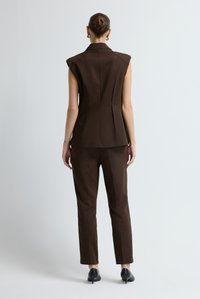 Frau mit einem Dutt, die eine ärmellose braune Blazer-Jacke, passende Hose, goldene Creolen und schwarze Kitten-Heels trägt, steht vor einer weißen Wand.