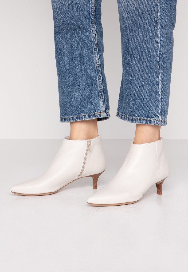 Bottines blanches en cuir avec fermeture éclair latérale et petits talons en bois, portées avec un jean bleu raccourci.