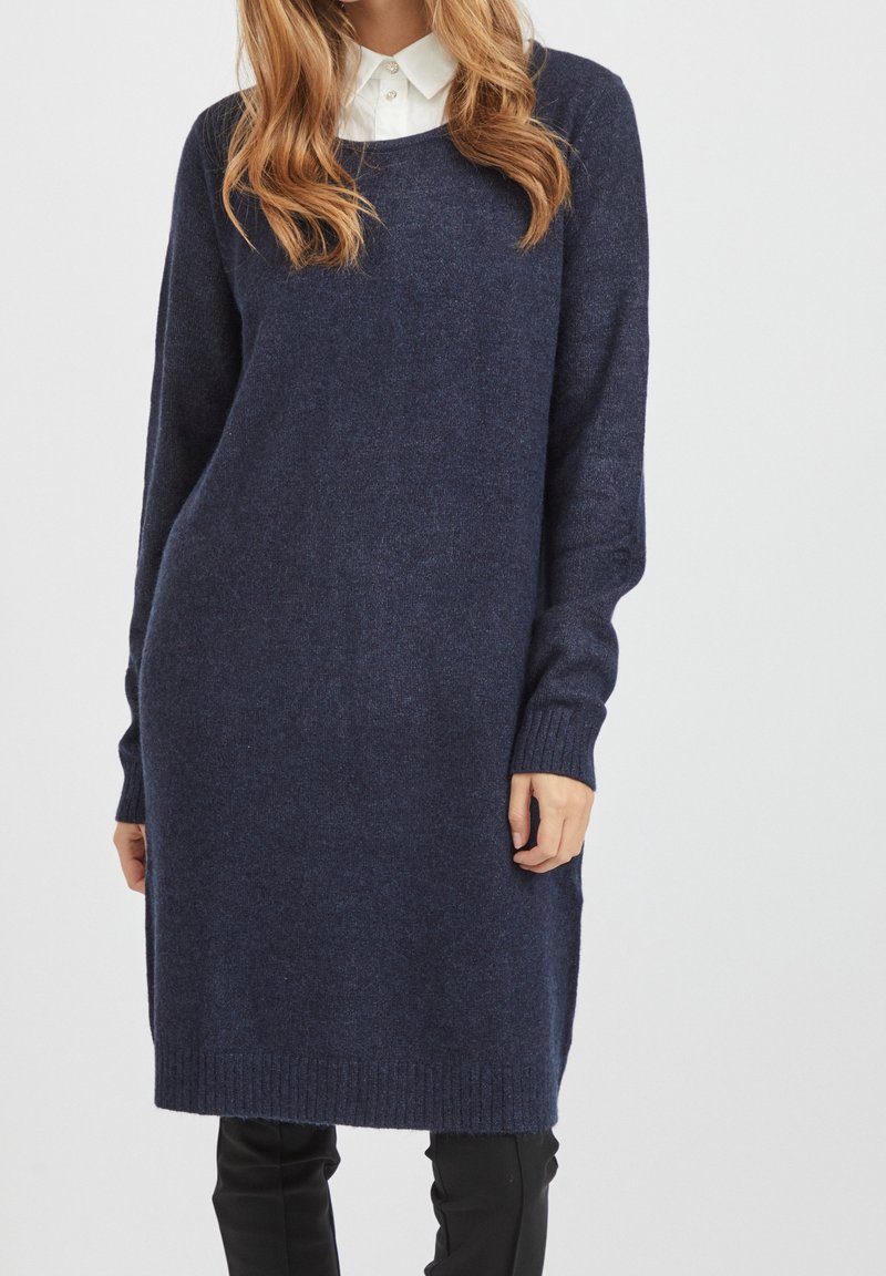 Femme portant une longue robe pull en maille bleu marine sur une chemise blanche à col et un pantalon noir, debout devant un fond uni.