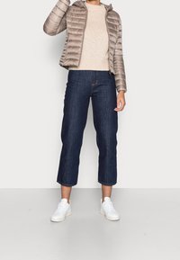 Beżowa pikowana kurtka z kapturem, sweter o ribbeyondanym splocie, ciemnoniebieskie, ścięte dżinsy oraz białe trampki, stojąca na jasnej podłodze na tle białej ściany.