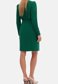 Robe verte à manches longues, taille ceinturée, et accents de boutons sur les poignets, fabriquée en tissu lisse. Portée avec des chaussures à talons de couleur claire.