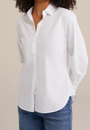 Kobieta w białej koszuli zapinanej na guziki z długim rękawem i niebieskich dżinsach, stojąca z rękami za plecami na jednolitym tle.