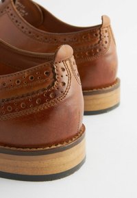 Zapatos de cuero marrón con un diseño de brogue, que presentan detalles perforados y acentos de costura. Tacón de madera con una base de goma negra.